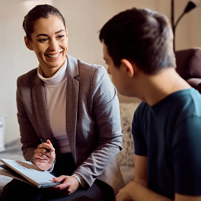 Smiling professional woman holding a notepad and pen while engaging in a supportive conversation with a young man in a casual setting.