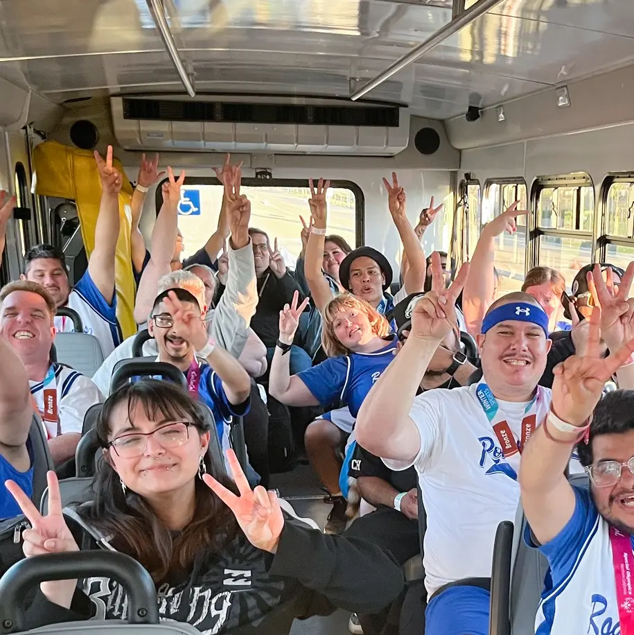 Group of smiling individuals on a bus, many wearing matching team shirts and medals, enthusiastically holding up peace signs. The group is celebrating as they return from a sports event, creating a joyful and inclusive atmosphere.