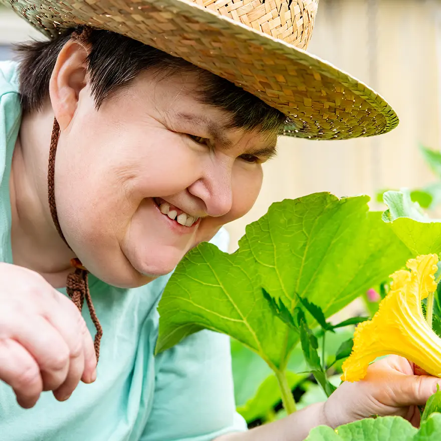 Smiling woman wearing a straw hat closely examining a vibrant yellow flower in a garden, surrounded by large green leaves. She appears joyful and engaged with nature.