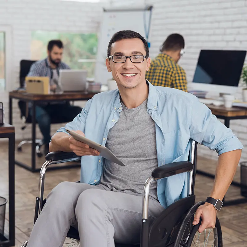 Smiling man in a wheelchair holding a tablet in a modern office setting, with coworkers working in the background at desks and computers.