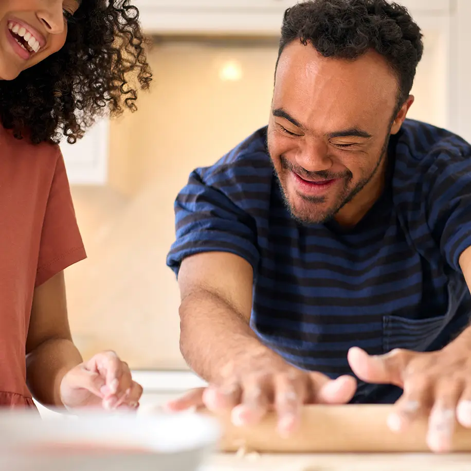 Smiling man rolling dough with a wooden rolling pin alongside a woman in a kitchen. Both appear joyful and engaged in a shared cooking activity.