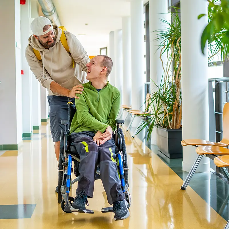 Support worker gently pushing a smiling man in a wheelchair down a bright hallway lined with plants and chairs. They are making eye contact, sharing a joyful and caring moment.