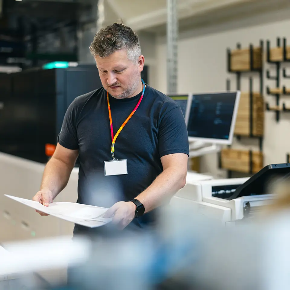 A man wearing a name badge is examining printed documents in a print shop, standing beside high-tech printing equipment and computer monitors.