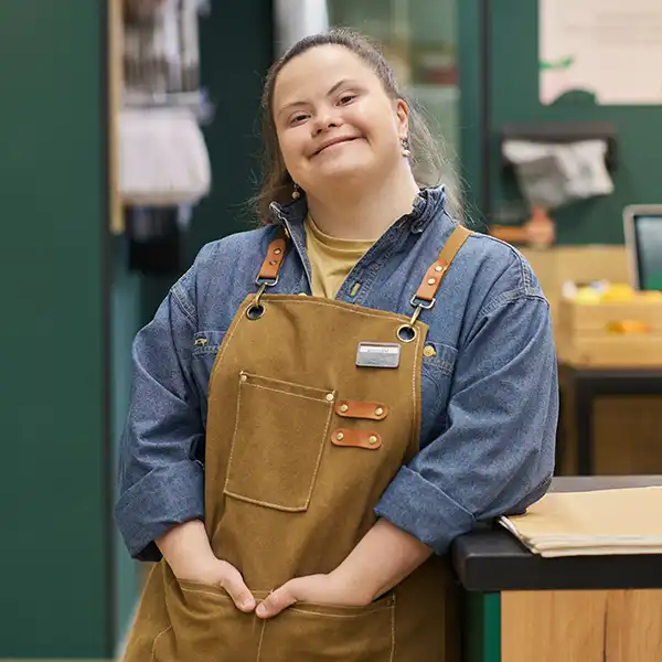 Smiling woman wearing a denim shirt and brown apron standing confidently behind a counter in a retail or service environment, hands in her pockets.