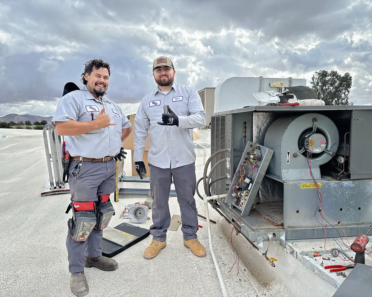 Two PSW maintenance team members stand on a rooftop next to HVAC equipment, smiling and giving hand gestures while working under a cloudy sky. Both wear PSW uniforms and safety gear, with tools and parts laid out around them.