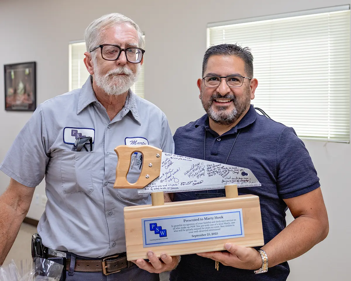 Marty Houk stands beside a smiling colleague at his retirement celebration, holding a custom wooden saw-shaped award engraved with messages from coworkers and a PSW plaque recognizing his dedicated service.